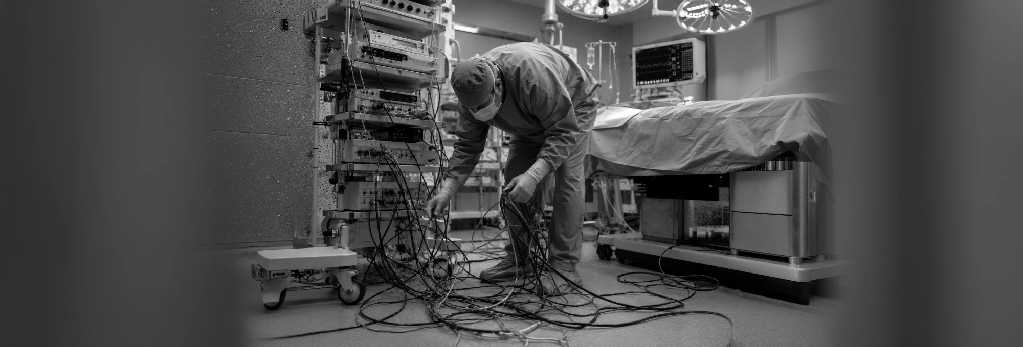 Clinician in scrubs untangling a mass of video cables on the floor beside an equipment tower in an operating room